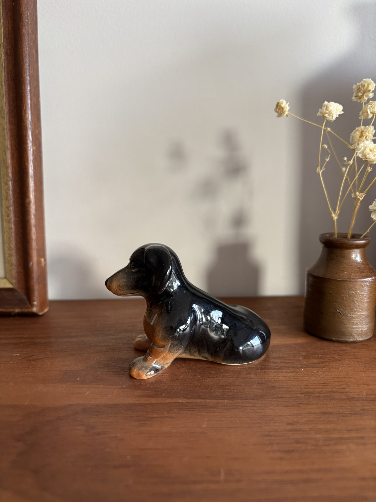 A glossy ceramic dachshund figurine in black and tan sits on a wooden table next to a small brown vase with dried flowers.