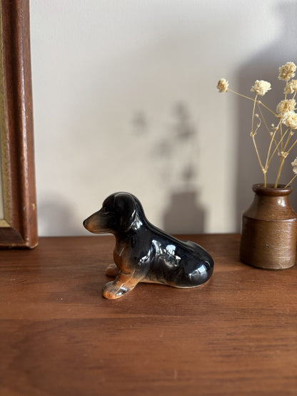 A glossy ceramic dachshund figurine in black and tan sits on a wooden table next to a small brown vase with dried flowers.