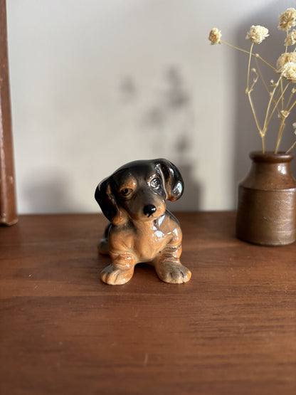 A small glossy ceramic dachshund figurine sitting on a wooden table, with a brown vase of dried flowers in the background.