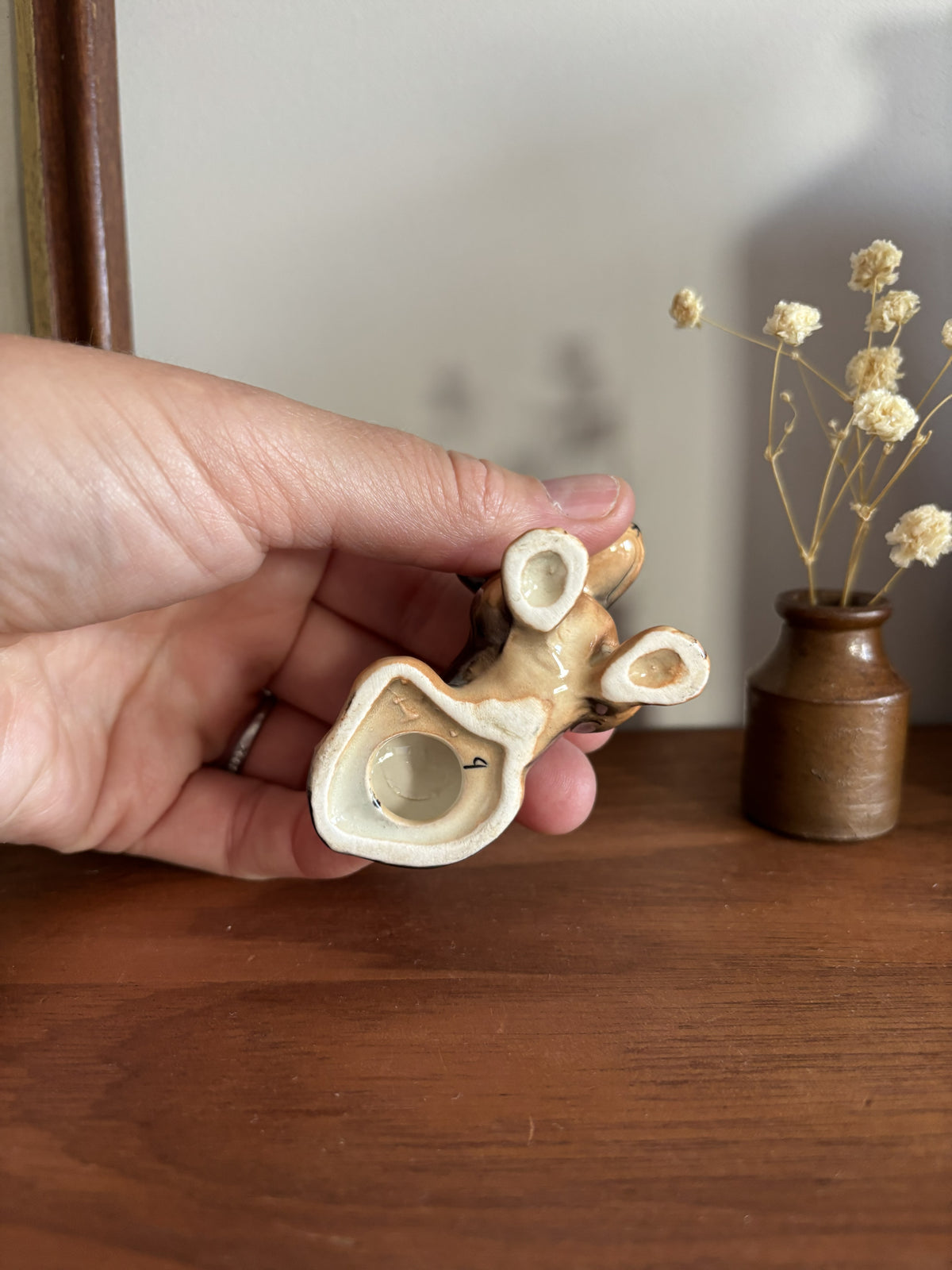 A hand holding a small ceramic Dachshund figurine, with a cream and brown glaze, set against a wooden table and a small vase of dried flowers.