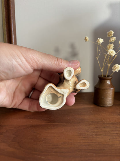 A hand holding a small ceramic Dachshund figurine, with a cream and brown glaze, set against a wooden table and a small vase of dried flowers.