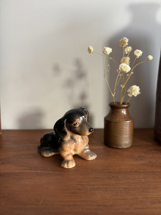 A small glossy ceramic dachshund figurine in black and tan sits on a wooden surface next to a rustic brown vase holding dried cream flowers, against a plain light wall.