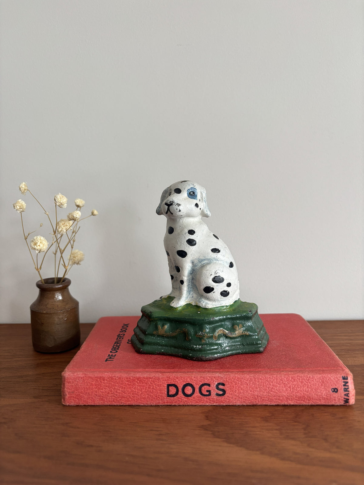 A cast iron Dalmatian door stop sitting on a green base, displayed on top of a red book on a wooden surface with a small vase of dried flowers to the left.