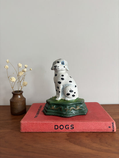 A cast iron Dalmatian door stop sitting on a green base, displayed on top of a red book on a wooden surface with a small vase of dried flowers to the left.