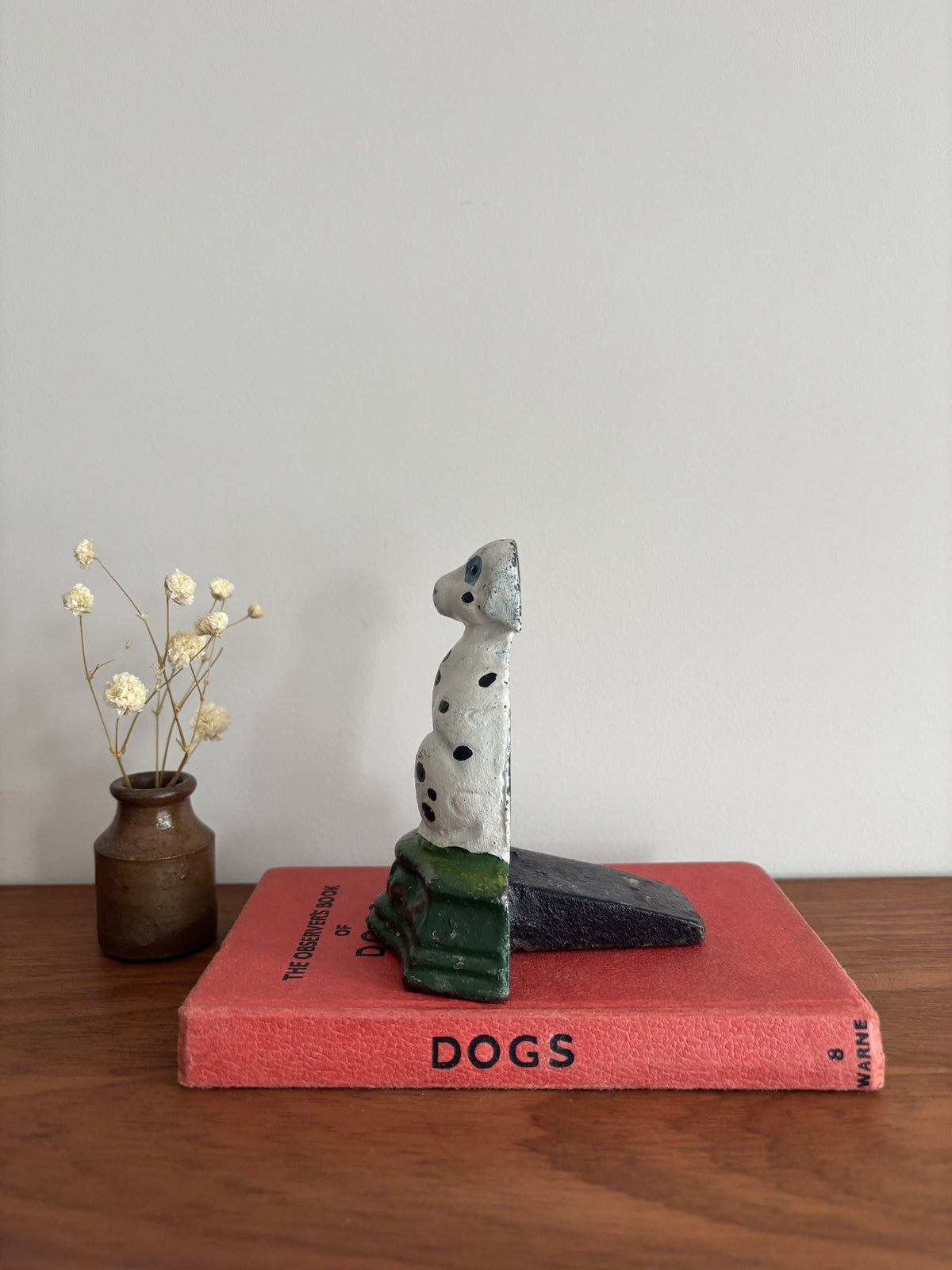 A cast iron Dalmatian door stop sitting on a green base, displayed on top of a red book on a wooden surface with a small vase of dried flowers to the left.