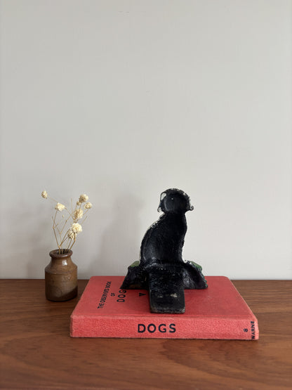 A cast iron Dalmatian door stop sitting on a green base, displayed on top of a red book on a wooden surface with a small vase of dried flowers to the left.