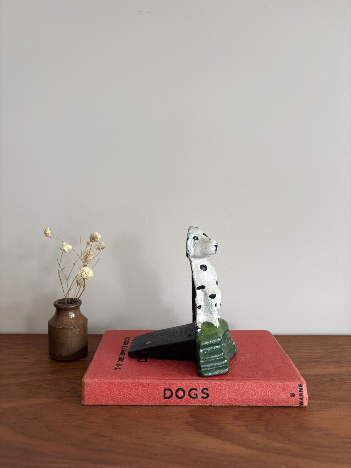 A cast iron Dalmatian door stop sitting on a green base, displayed on top of a red book on a wooden surface with a small vase of dried flowers to the left.
