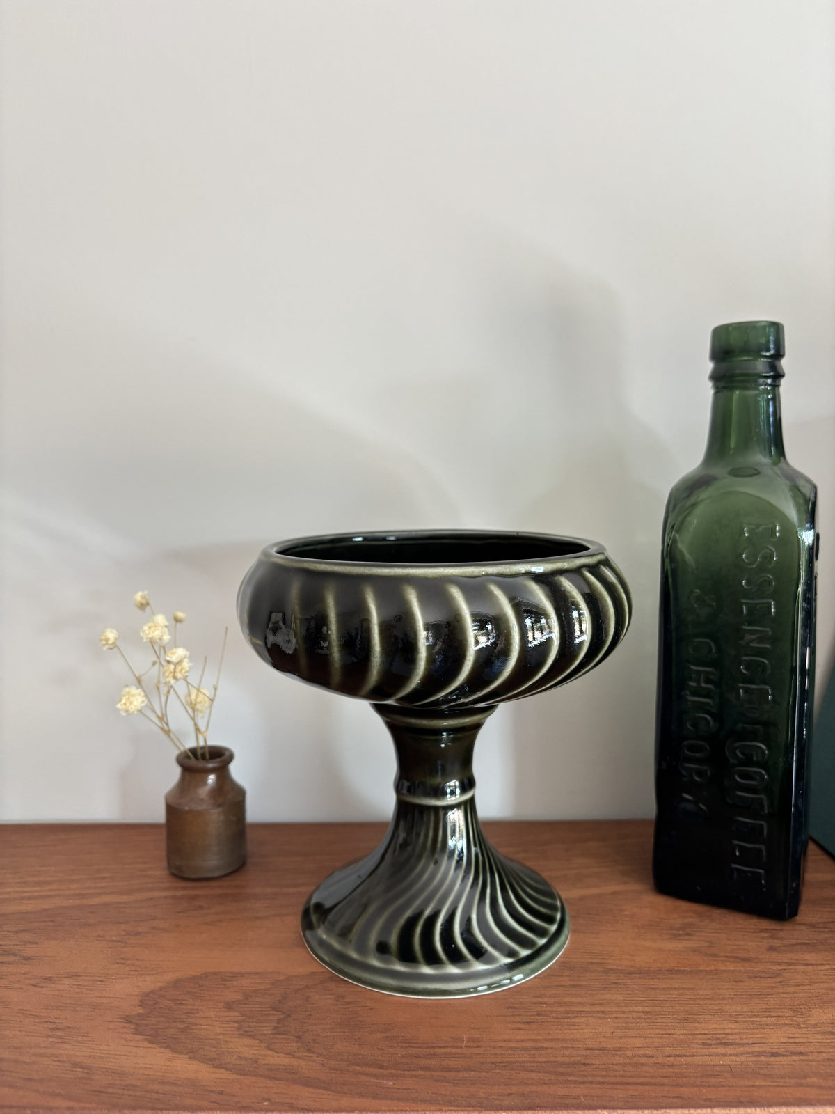 A dark green and black ribbed ceramic pedestal bowl with a flared base, displayed on a wooden table next to a vintage green glass bottle and a small vase with dried flowers.