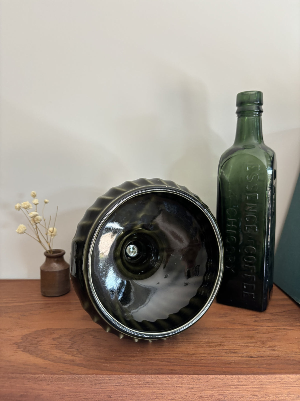 A dark green glass pedestal with a fluted rim  on a wooden surface next to a vintage green glass bottle and a small vase of dried flowers.