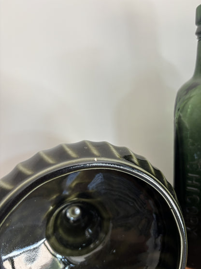 Close-up of a dark green glass pedestal lying on its side, showing the interior glass wall and neck, with another matching bottle partially visible on the right against a plain light background.