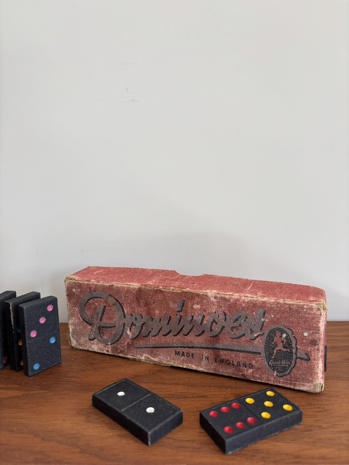 Vintage wooden domino game set with a worn red-brown box and black domino tiles on a wooden tabletop against a plain light wall.