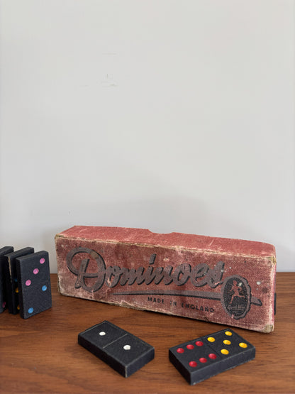 Vintage wooden domino game set with a worn red-brown box and black domino tiles on a wooden tabletop against a plain light wall.