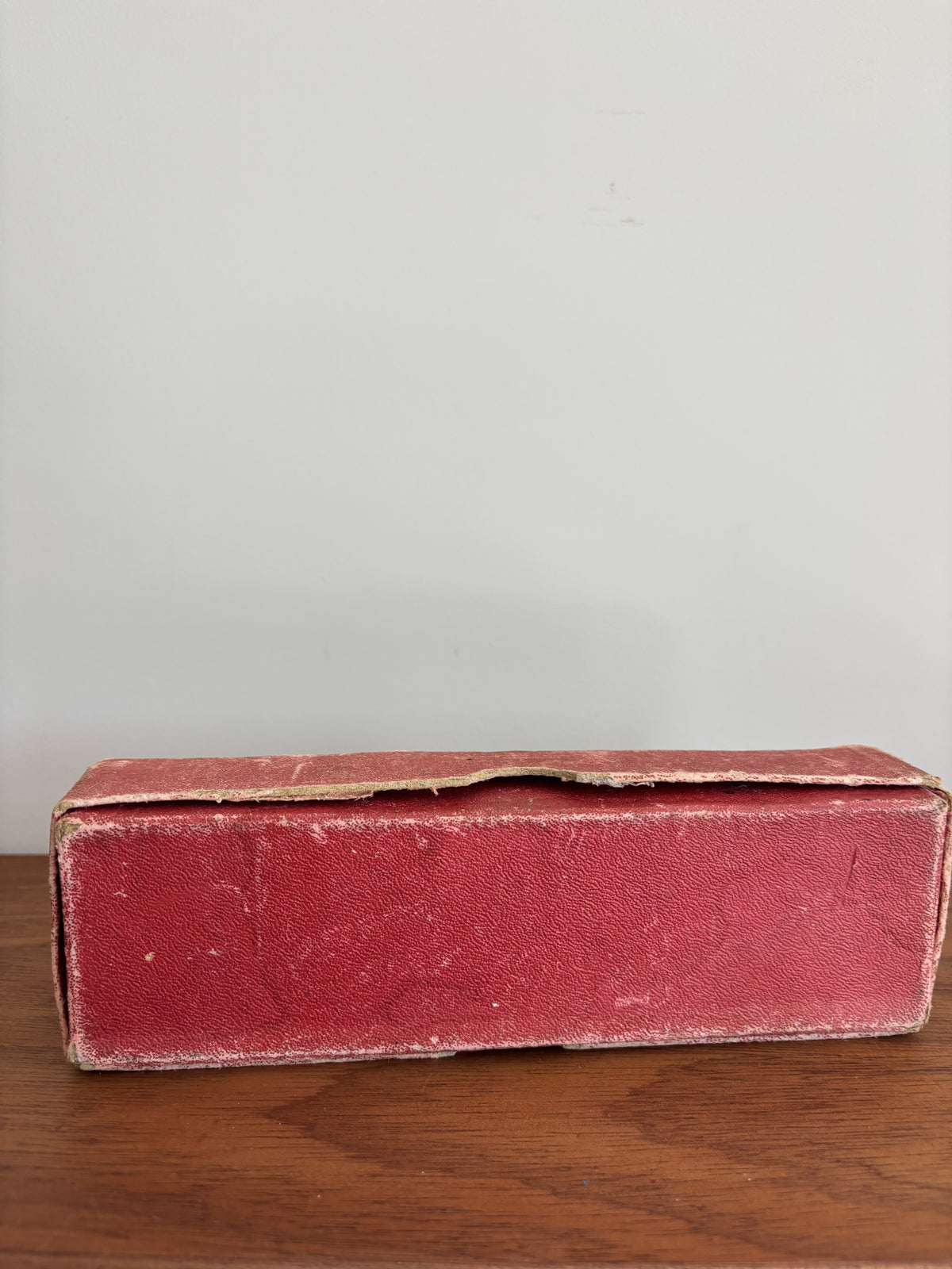 A vintage rectangular red box with a textured leather or faux-leather finish, resting on a wooden surface against a plain light gray wall.