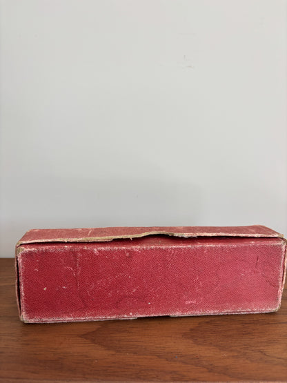 A vintage rectangular red box with a textured leather or faux-leather finish, resting on a wooden surface against a plain light gray wall.