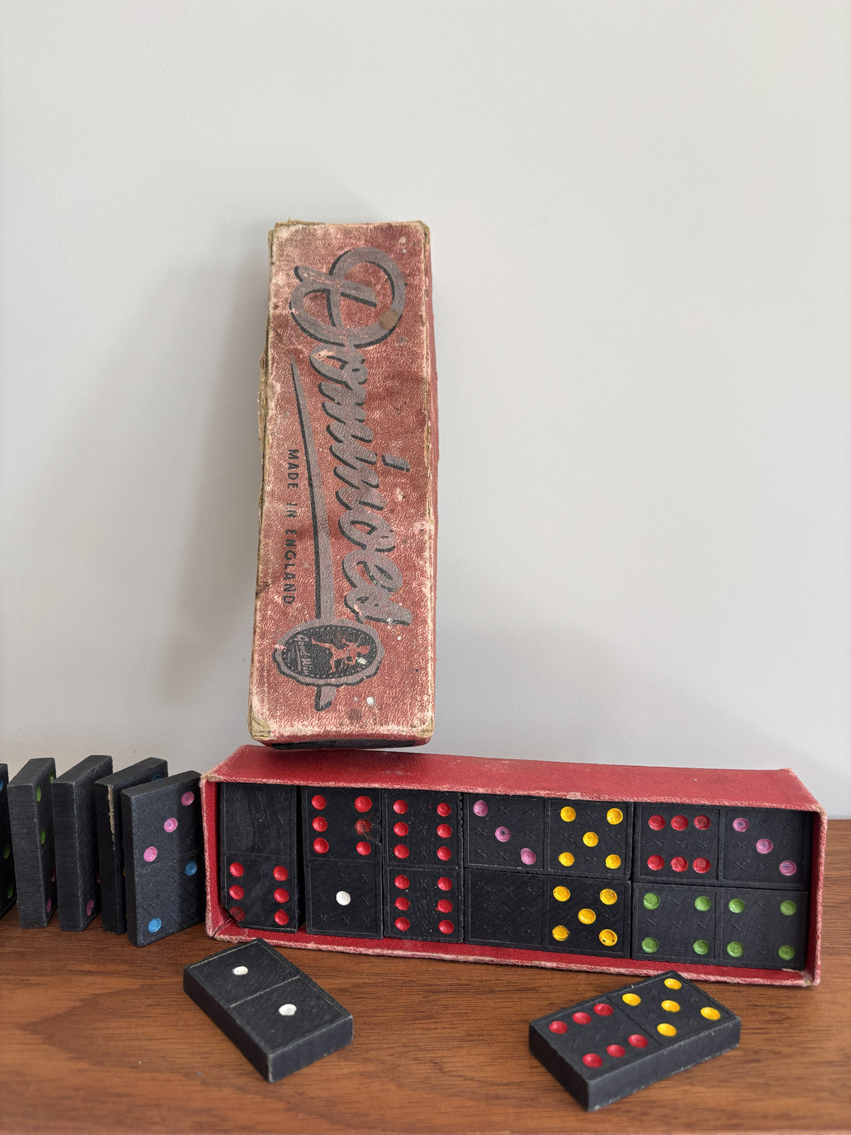 Vintage domino game set with a worn red box, black domino tiles, and colorful dot accents on a wooden surface against a plain light wall.