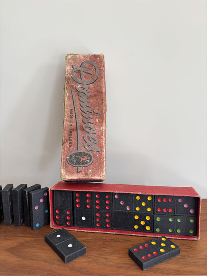 Vintage domino game set with a worn red box, black domino tiles, and colorful dot accents on a wooden surface against a plain light wall.