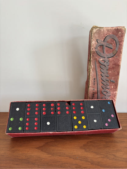Vintage domino game set with black domino tiles and colorful pips in a red box, shown next to its worn cardboard box on a wooden surface against a plain wall.