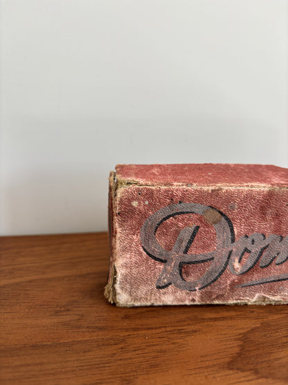 A close-up of a vintage, worn reddish-brown vintage domino box resting on a wooden tabletop against a plain light gray wall.