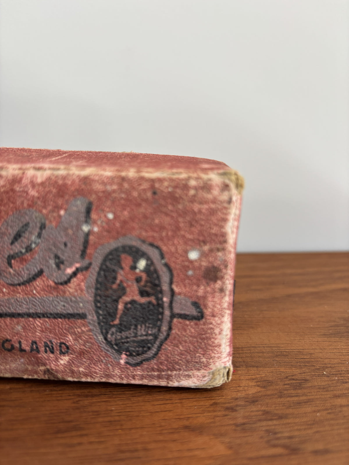 A close-up of a vintage, worn red box with black printed branding, resting on a wooden surface against a light gray background.