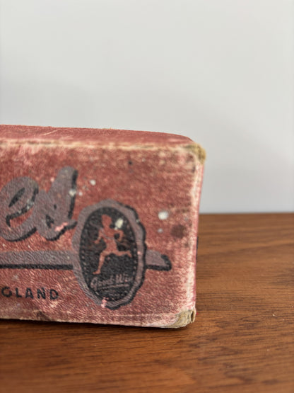 A close-up of a vintage, worn red box with black printed branding, resting on a wooden surface against a light gray background.