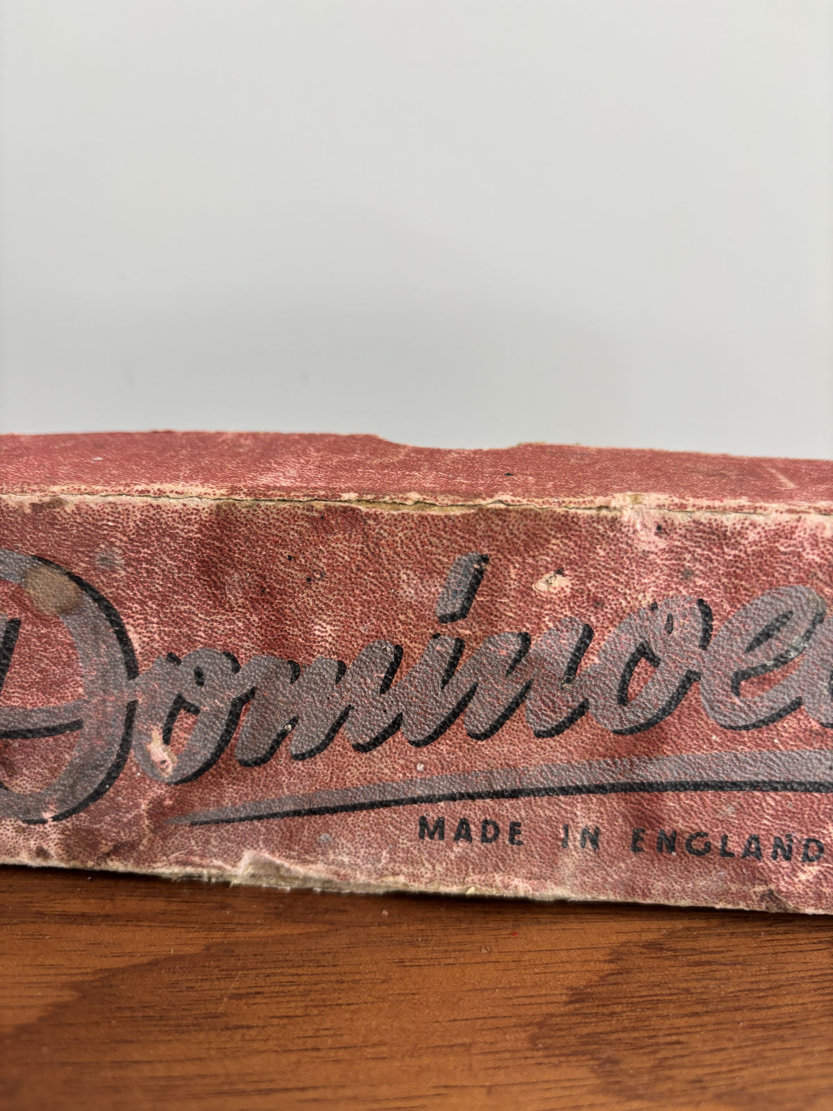 A close-up of a vintage red worn box with black 'Dominoes' logo and 'MADE IN ENGLAND' text, resting on a wooden surface against a light gray background.