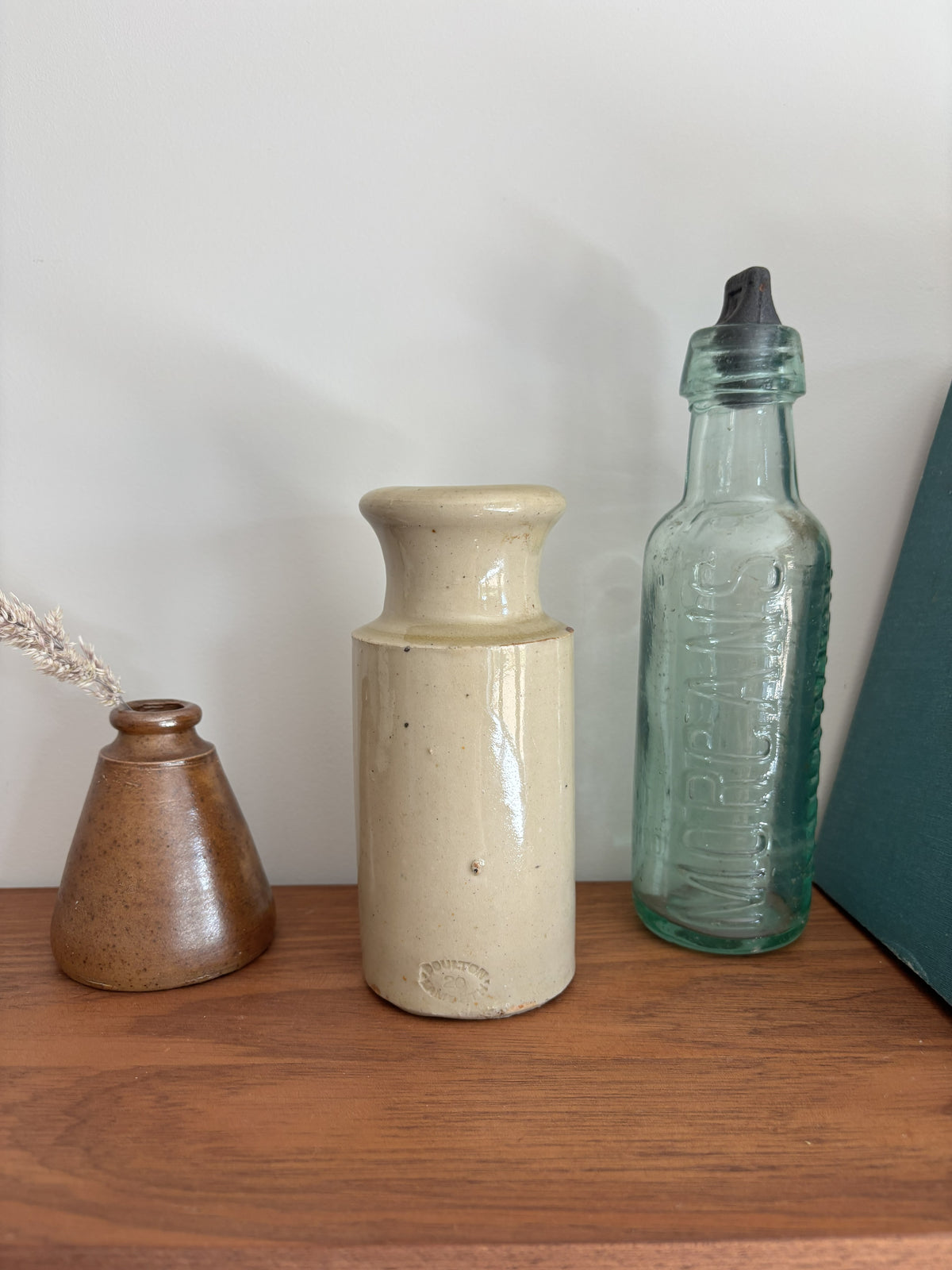 Vintage cream stoneware ink bottle displayed on a wooden shelf against a plain light gray wall styled with a vase with dried grass and a tall translucent green glass bottle with a black cap.