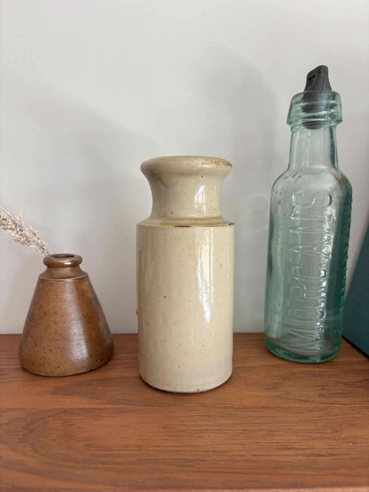 Vintage cream stoneware ink bottle displayed on a wooden shelf against a plain light gray wall styled with a vase with dried grass and a tall translucent green glass bottle with a black cap.