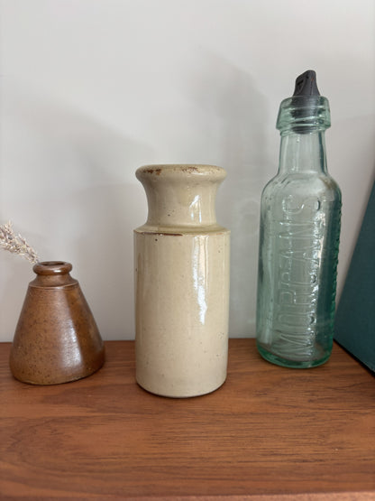 Vintage cream stoneware ink bottle displayed on a wooden shelf against a plain light gray wall styled with a vase with dried grass and a tall translucent green glass bottle with a black cap.