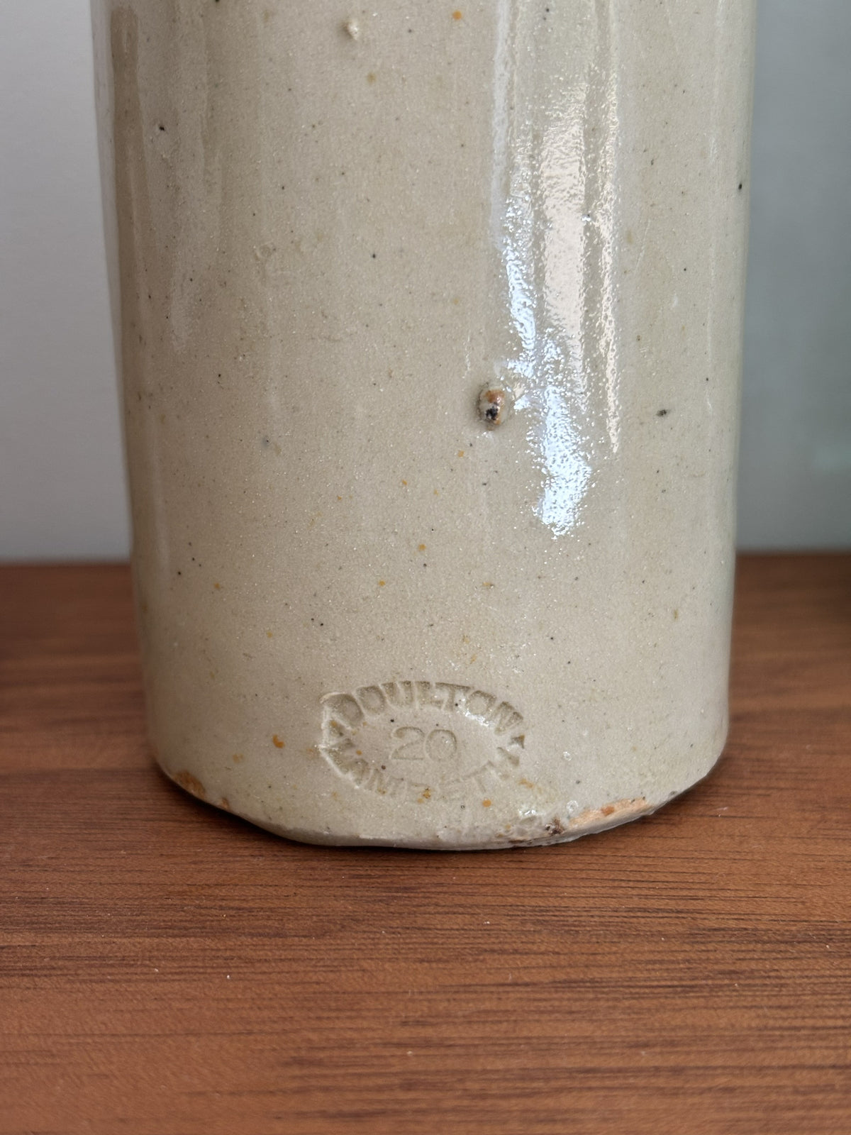 Close-up of a vintage cream stoneware bottle, showing a stamped 'Doulton Lambeth 20' mark on the bottom, resting on a wooden surface.