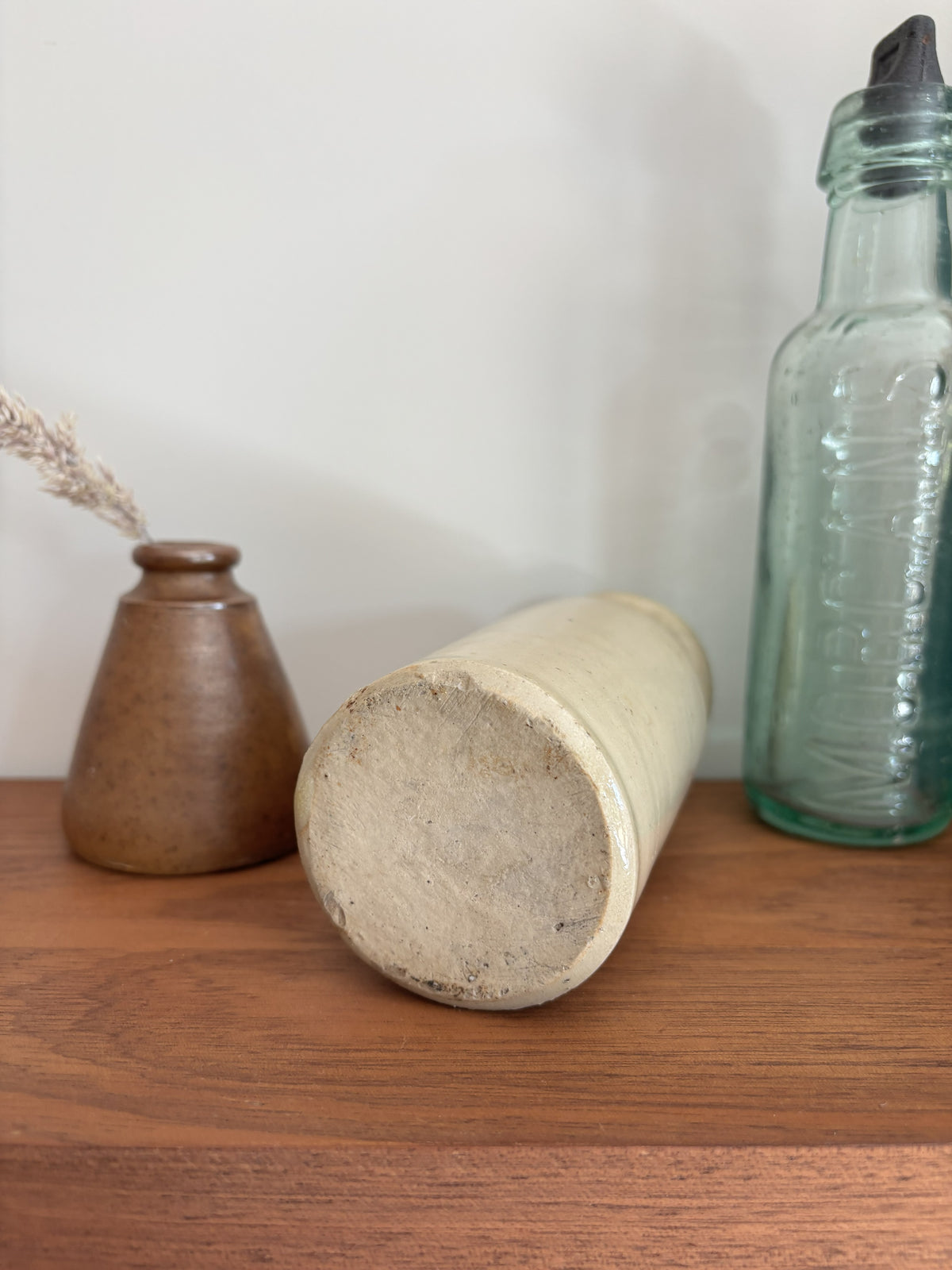 The base of a vintage cream stoneware ink bottle displayed on a wooden shelf against a plain light gray wall styled with a vase with dried grass and a tall translucent green glass bottle with a black cap.