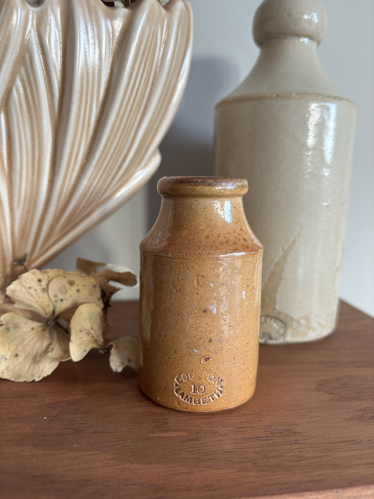 A small vintage stoneware bottle with a speckled brown glaze sits on a wooden surface, styled with dried flowers and other ceramic decor in the background.