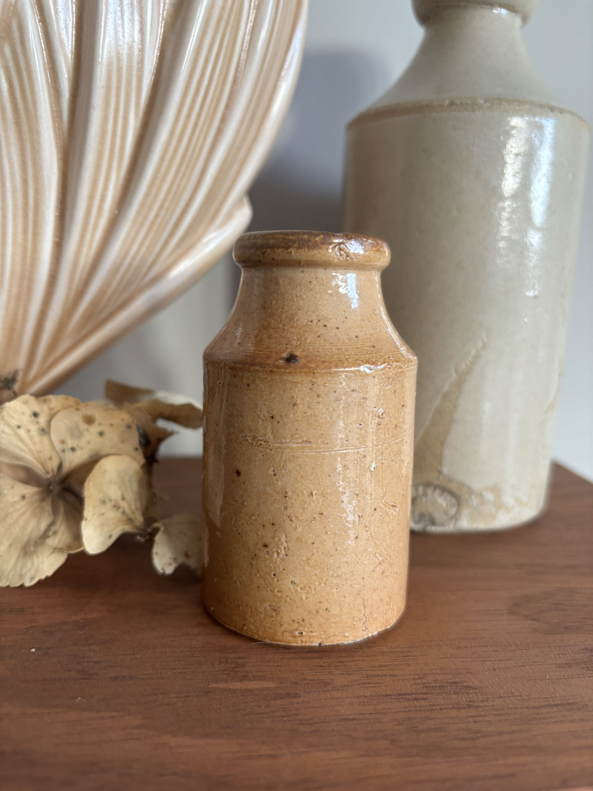 A small vintage stoneware bottle with a speckled tan-brown glaze sits on a wooden surface, styled with dried flowers and other ceramic vases in the background.
