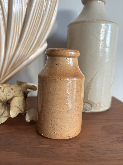 A small vintage stoneware bottle with a warm tan-brown glaze sits on a wooden surface, styled with dried flowers and a larger cream-colored vase in the background.