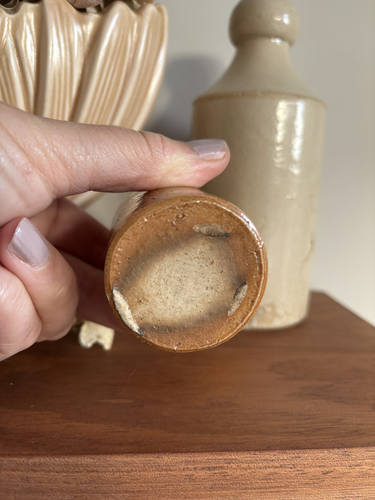 A close-up of a hand holding the bottom of a small brown stoneware bottle showing the glazed base. A larger beige ceramic bottle or vase stands in the background on a wooden surface.