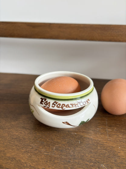 A white ceramic egg separator with brown text reading 'Egg Separator' sits on a wooden table, holding a brown egg while another egg rests beside it.
