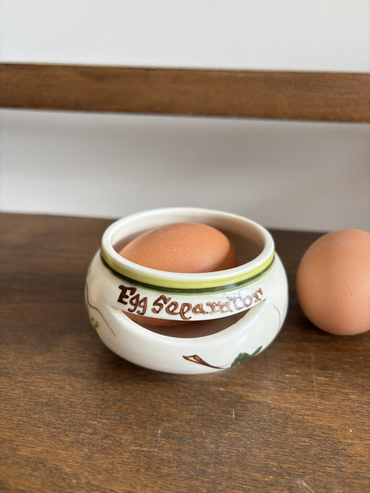 A white ceramic egg separator with brown text reading 'Egg Separator' sits on a wooden table, holding a brown egg while another egg rests beside it.