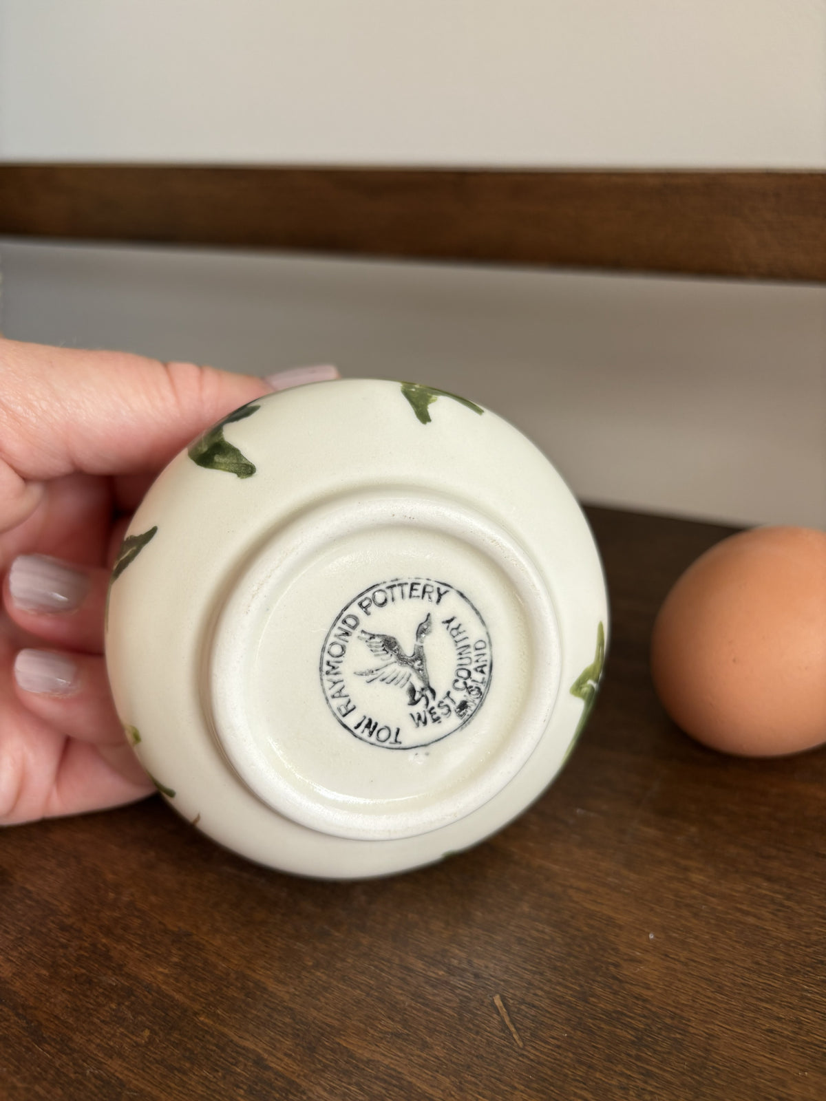 A hand holds the bottom of a small white ceramic bowl or cup with green leaf-like painted details, showing a printed potter's mark. A brown egg sits on a wooden surface in the background.