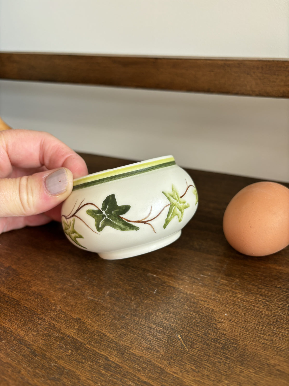 A hand holding a small white ceramic egg cup decorated with green ivy leaves and brown vines, with a brown egg on a wooden table next to it.