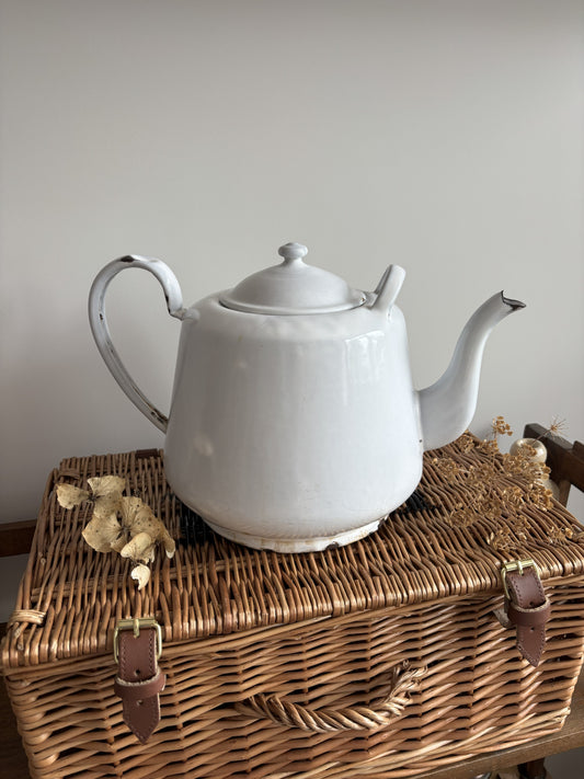 A white enamel teapot with a rounded body, curved handle, and spout, displayed on top of a brown woven wicker trunk against a plain light gray wall.