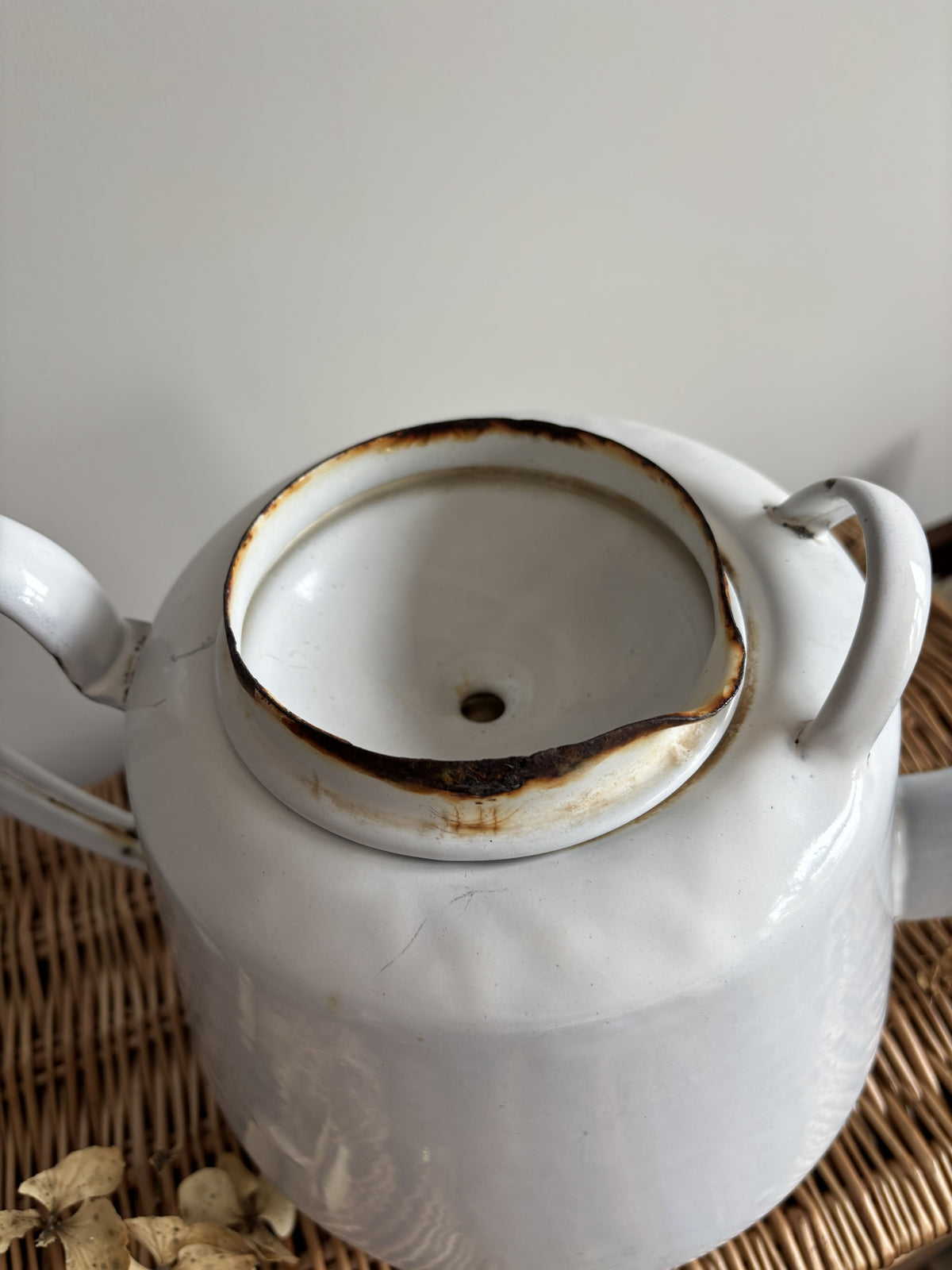 A white enamel teapot with a rounded body, curved handle, and spout, displayed on top of a brown woven wicker trunk against a plain light gray wall.