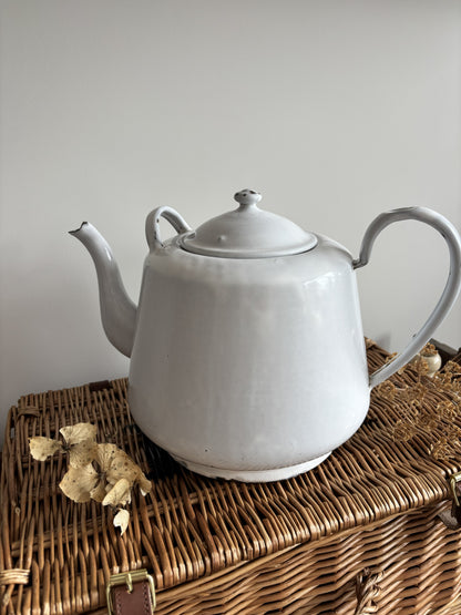 A white enamel teapot with a rounded body, curved handle, and spout, displayed on top of a brown woven wicker trunk against a plain light gray wall.