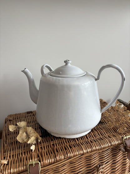 A white enamel teapot with a rounded body, curved handle, and spout, displayed on top of a brown woven wicker trunk against a plain light gray wall.