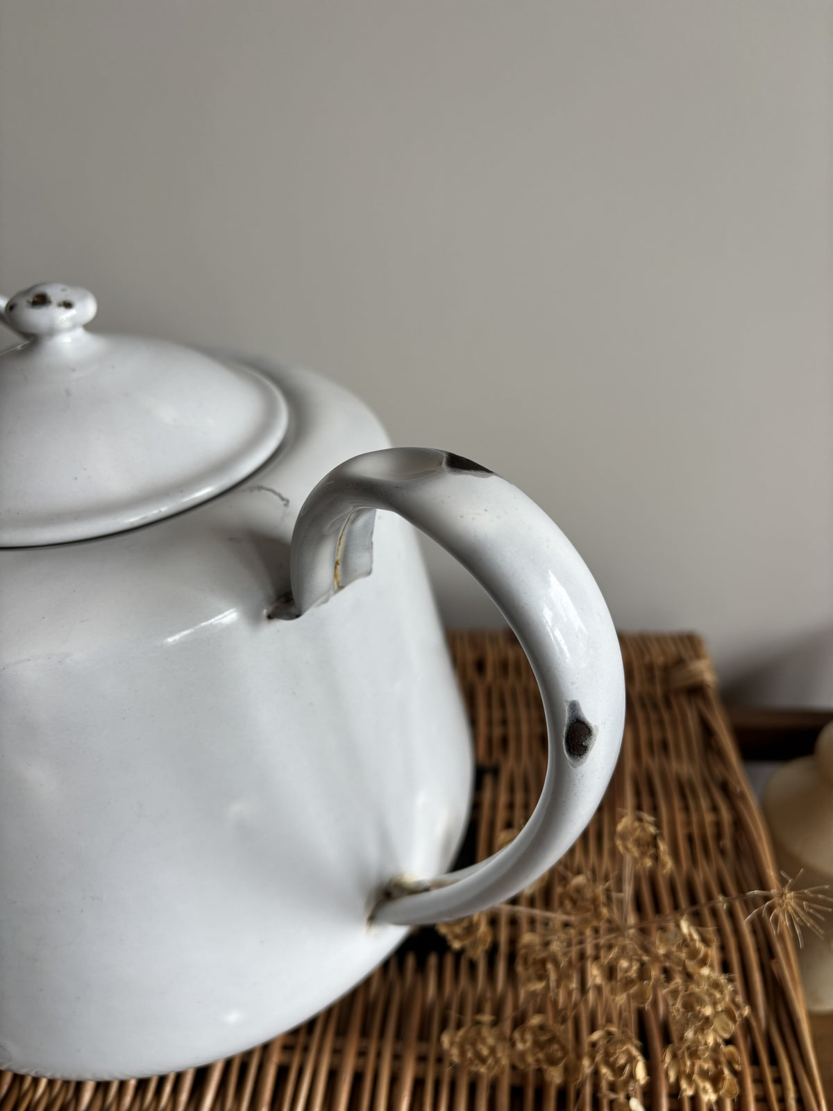 A white enamel teapot with a rounded body, curved handle, and spout, displayed on top of a brown woven wicker trunk against a plain light gray wall. Close up of chips on handle