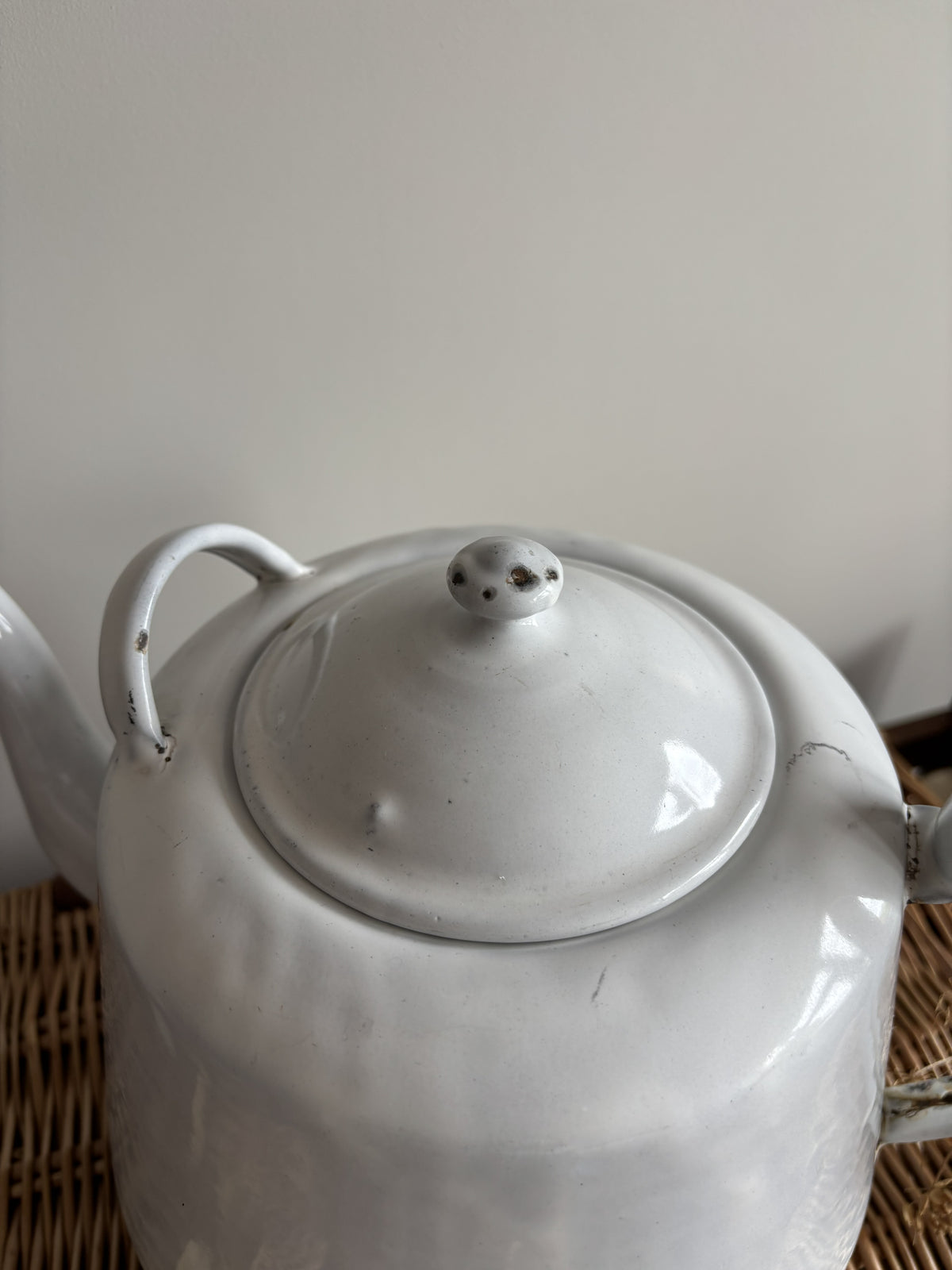 A white enamel teapot with a rounded body, curved handle, and spout, displayed on top of a brown woven wicker trunk against a plain light gray wall.