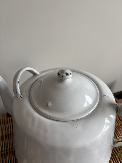 A white enamel teapot with a rounded body, curved handle, and spout, displayed on top of a brown woven wicker trunk against a plain light gray wall.