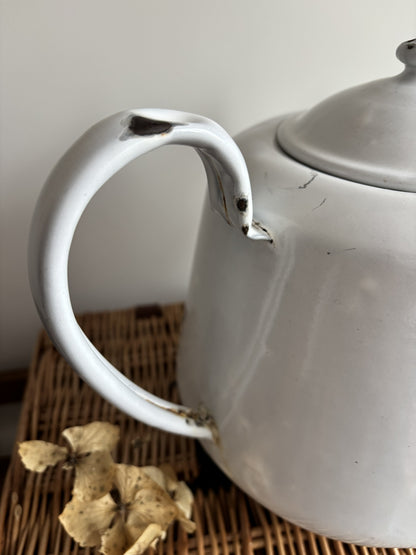 A white enamel teapot with a rounded body, curved handle, and spout, displayed on top of a brown woven wicker trunk against a plain light gray wall.