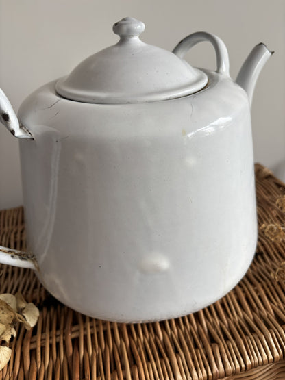A white enamel teapot with a rounded body, curved handle, and spout, displayed on top of a brown woven wicker trunk against a plain light gray wall.