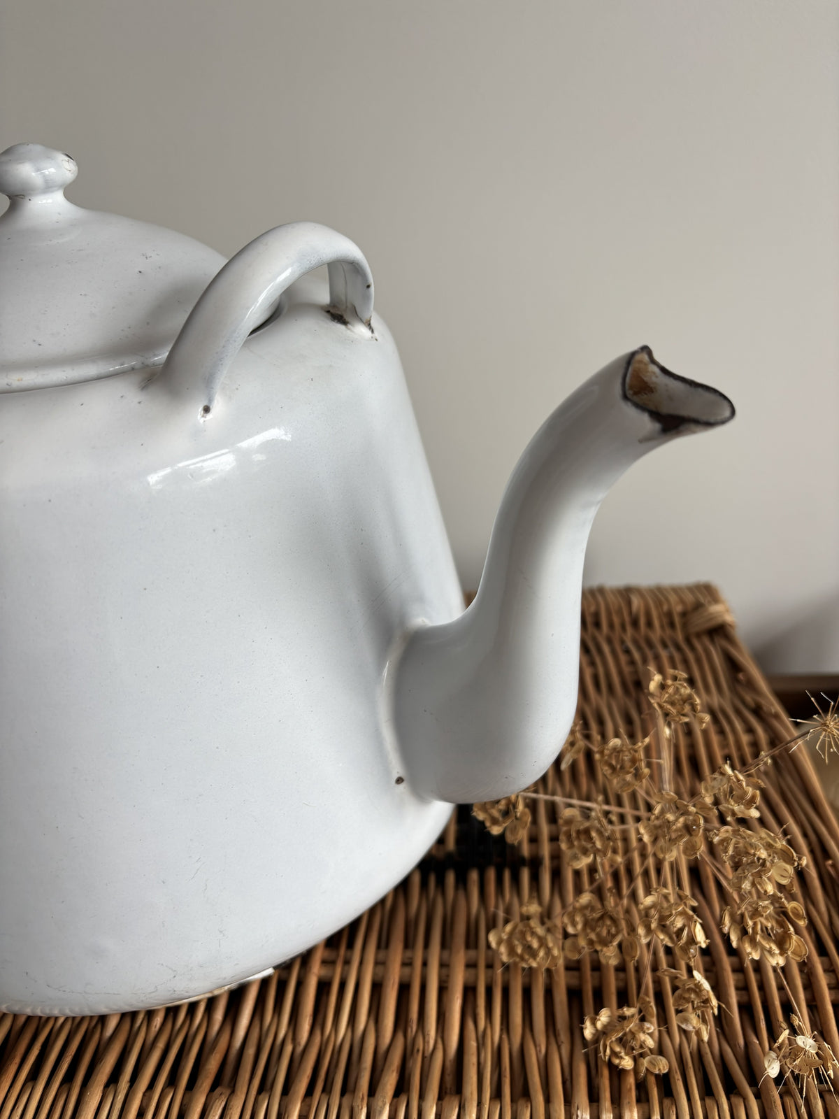 A white enamel teapot with a rounded body, curved handle, and spout, displayed on top of a brown woven wicker trunk against a plain light gray wall.