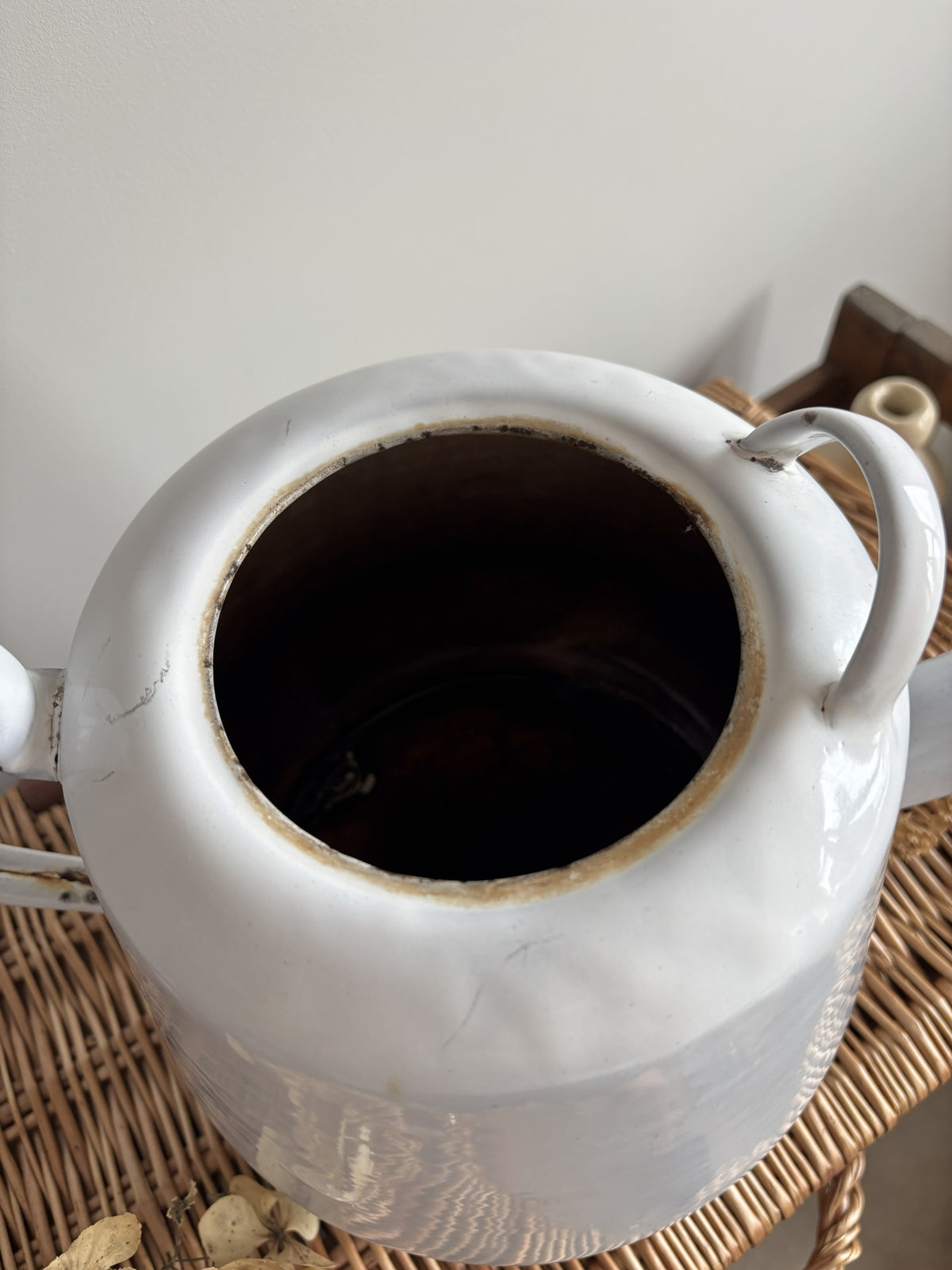 A white enamel teapot with a rounded body, curved handle, and spout, displayed on top of a brown woven wicker trunk against a plain light gray wall. Close up of inside.  
