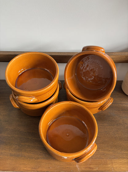 Five small glazed ceramic bowls with side handles, stacked and arranged on a wooden surface against a light wall.